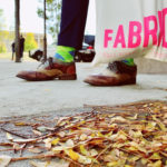 A pair of feet, holding a Fabric Festival tote bag on a sidewalk 