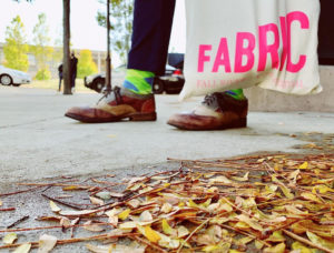 A pair of feet, holding a Fabric Festival tote bag on a sidewalk